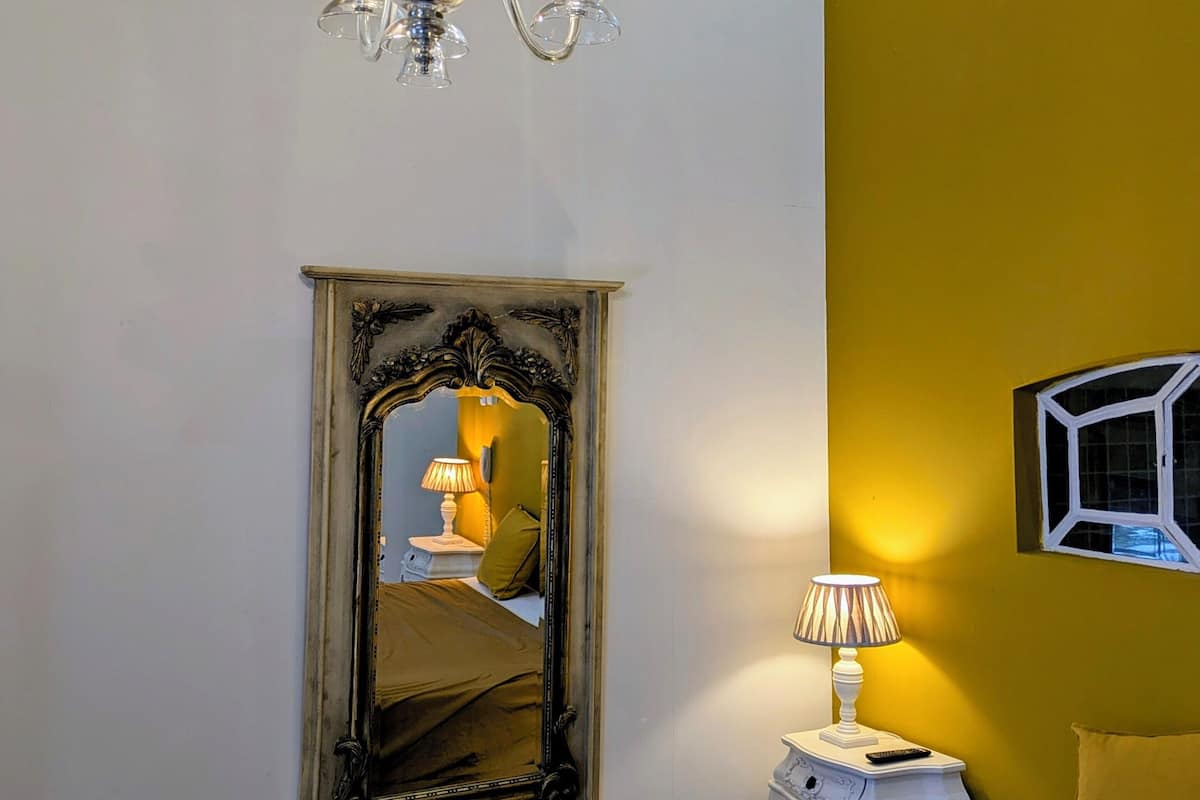 A bedroom with a bed, a chandelier, a mirror, a bedside table with a lamp, and a window with a view.