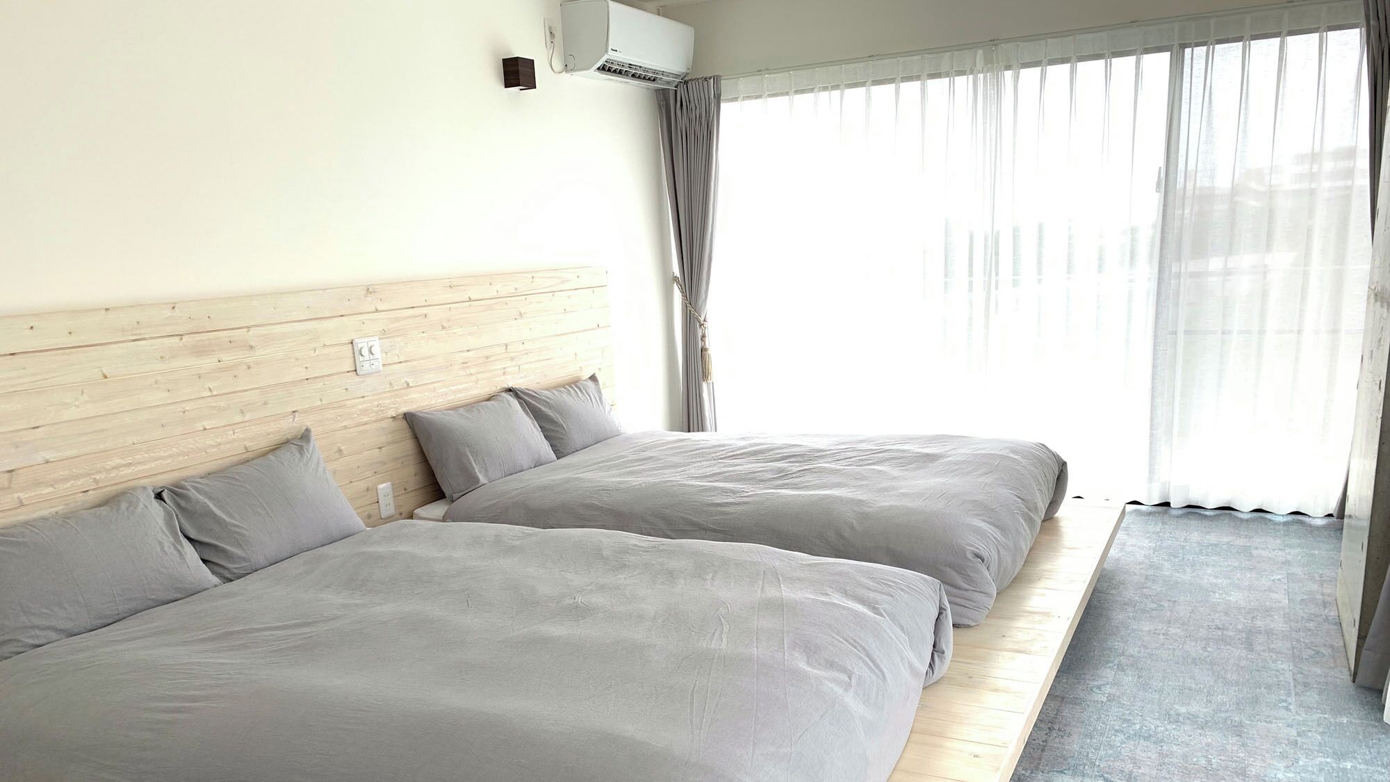 A hotel room with two beds, a wooden headboard, an air conditioning unit, and a large window with sheer curtains.