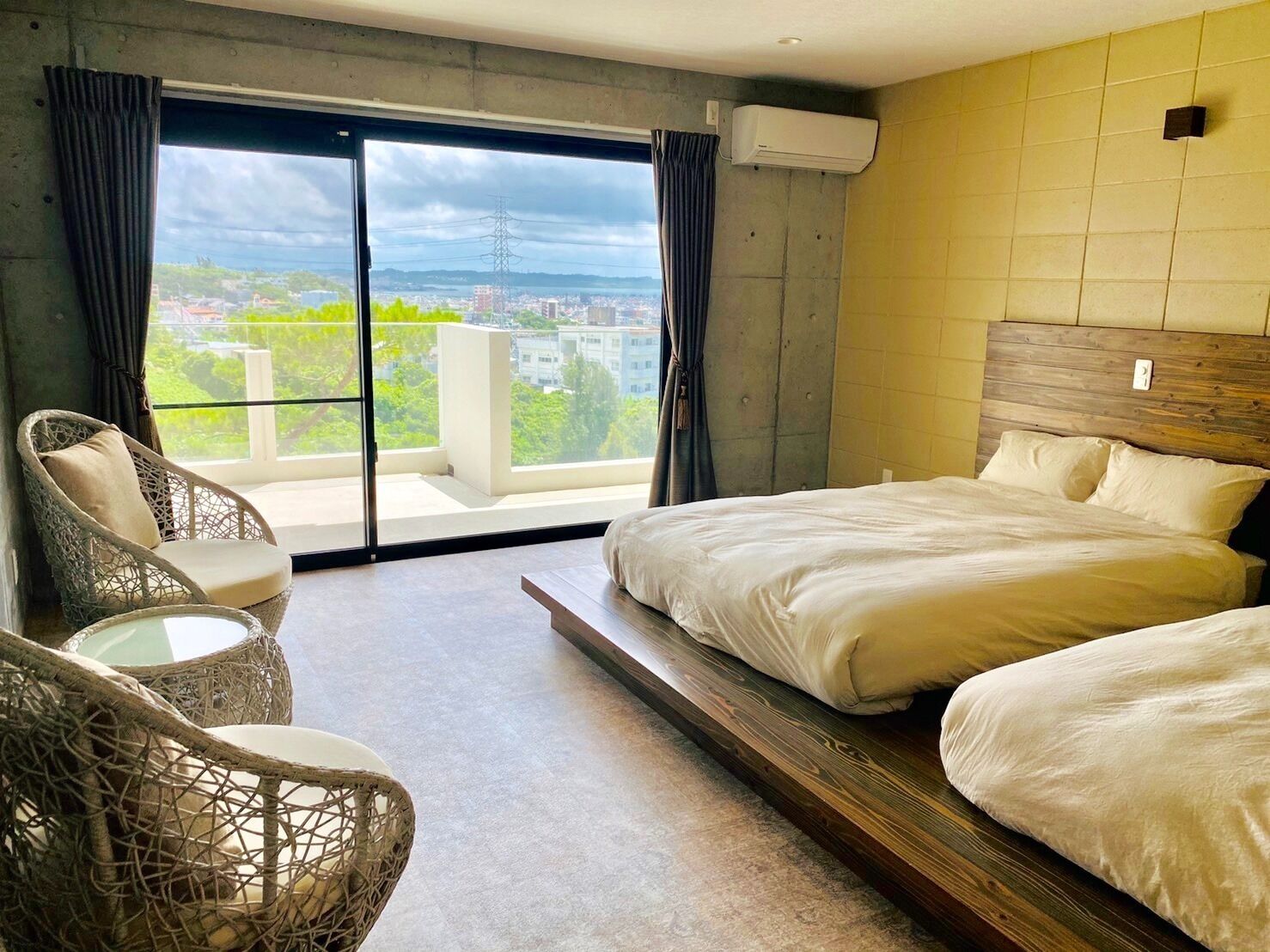 A hotel room with a bed, a chair, a small table, and a view of the city through large windows.