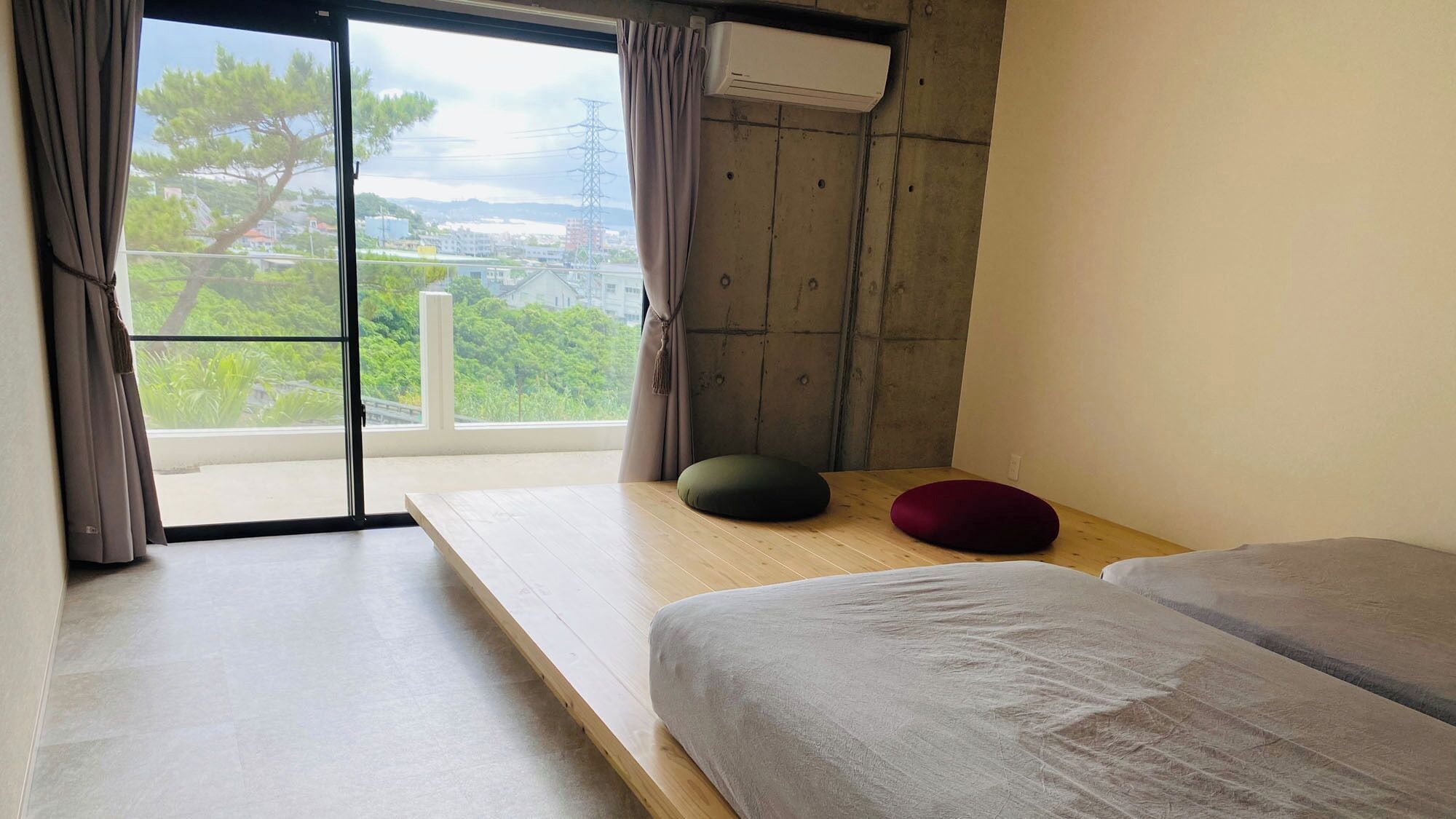 A room with a large window, a wooden floor, a bed with a grey blanket, a green and a red cushion, and a wall-mounted air conditioner.