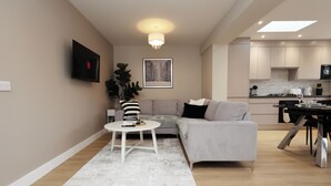 Ein modernes Wohnzimmer mit einem grauen Sofa, einem kleinen weißen Tisch mit einem Buch und einer großen Pflanze. Im Hintergrund ist eine Küche mit modernen Geräten zu sehen.