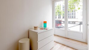 Ein schlicht eingerichtetes Zimmer mit einer weißen Kommode, einer kleinen Mülltonne und einem Fenster mit Blick auf ein Gebäude und Bäume.