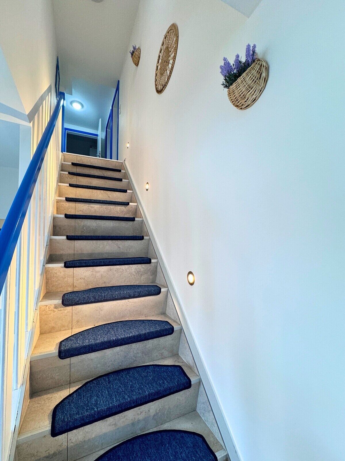 A staircase with beige steps and blue carpet runners, leading to a landing with a white wall and a basket with purple flowers.