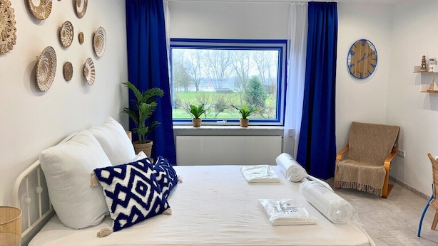 Hotellihuone, jossa on sänky, tuoli, ikkuna verhoilla, kello ja seinäkoristeita.