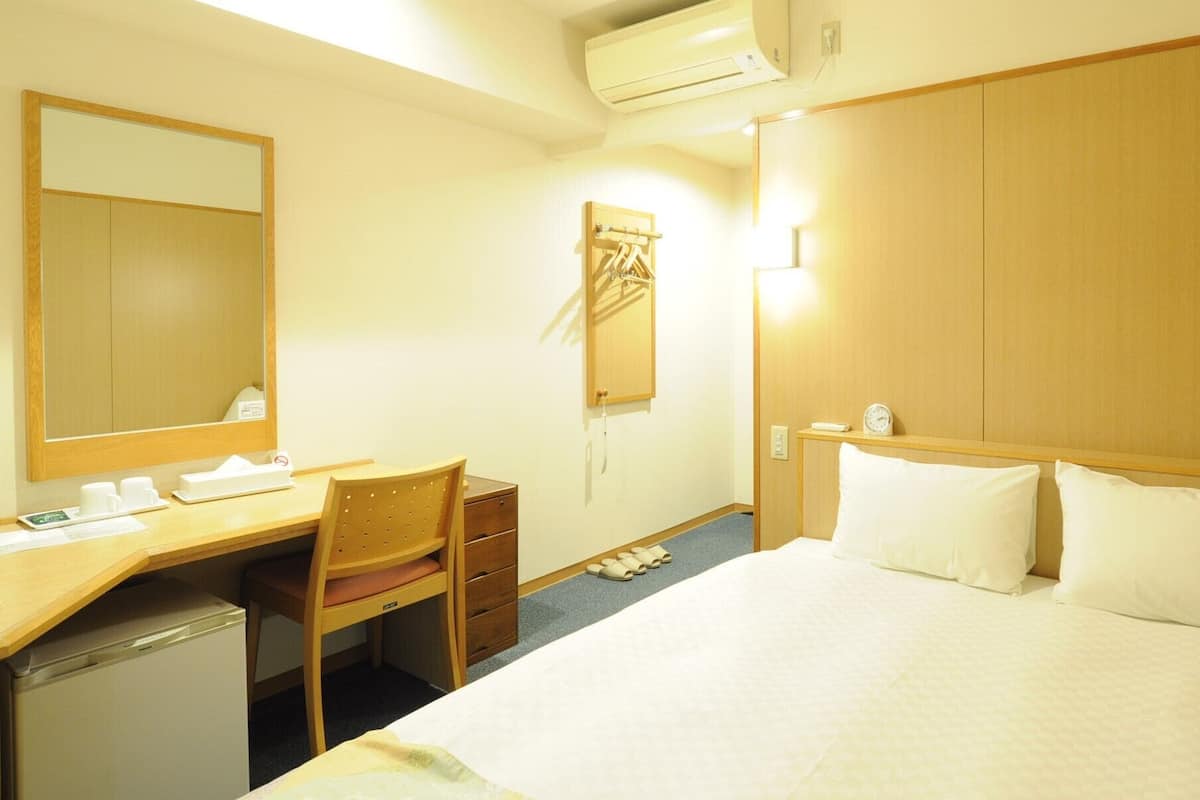 A hotel room with a bed, a desk, a chair, a mirror, a wardrobe, and an air conditioning unit.