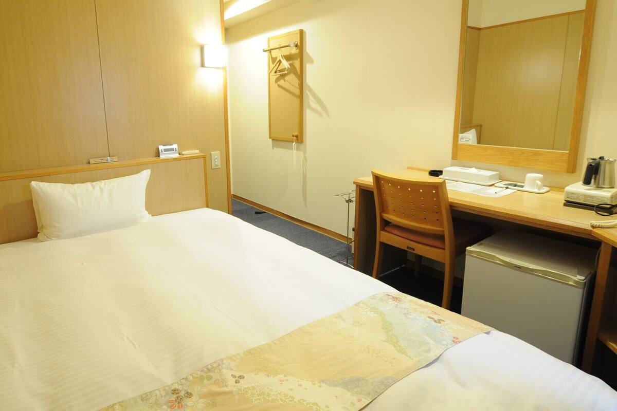 A hotel room with a bed, a desk, a chair, a mirror, and a small kitchenette.