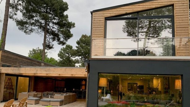 Una casa moderna de dos pisos amb un gran finestral, zona d'estar a l'exterior i un jardí ben cuidat.
