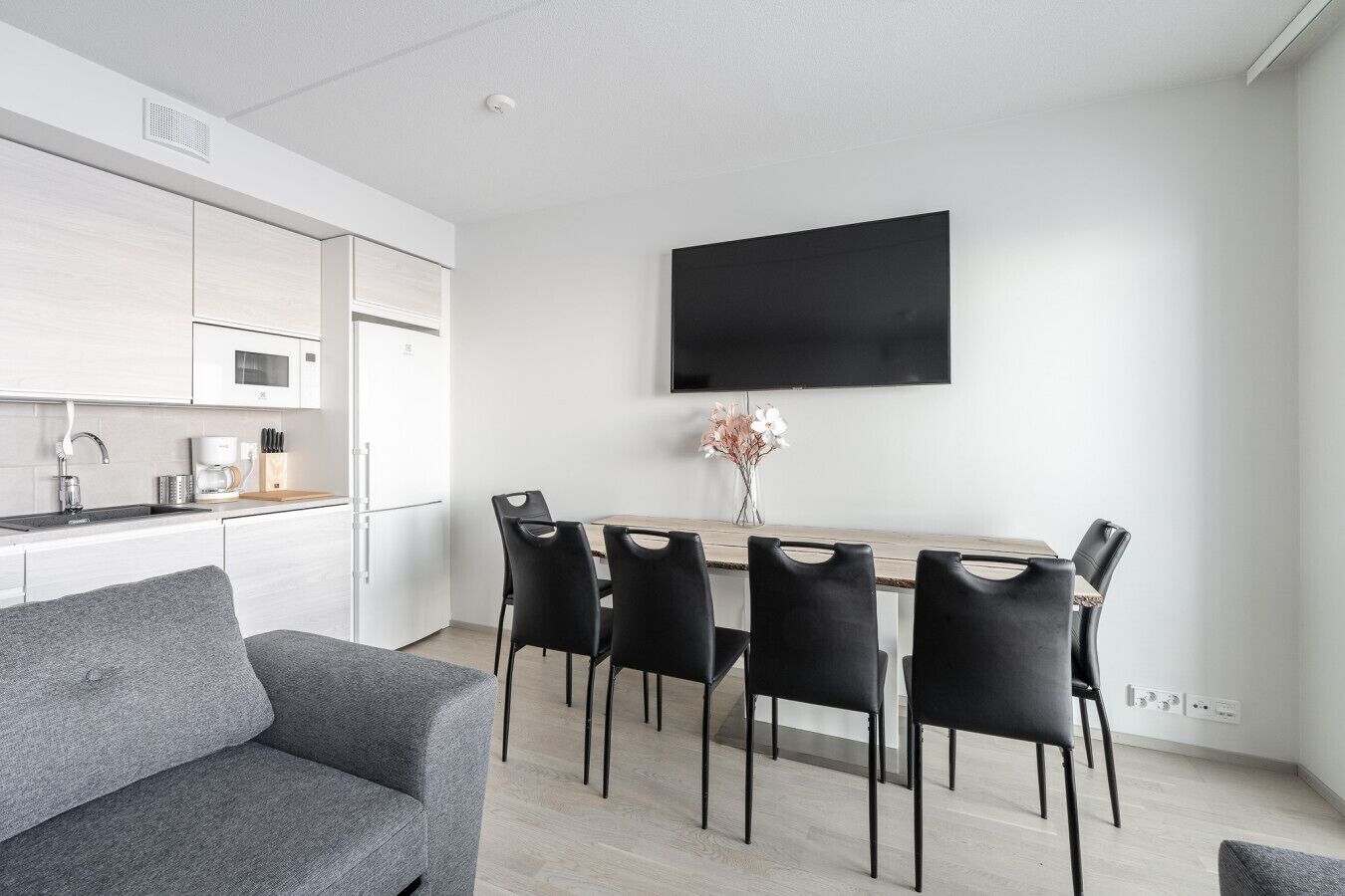 A modern kitchen with white cabinets, a built-in microwave, and a coffee machine. There is a dining area with a wooden table and black chairs. A flat-screen TV is mounted on the wall above the table.