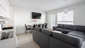 Ein modernes Wohnzimmer mit einem grauen Ecksofa, einem Essbereich mit Tisch und Stühlen, einem an der Wand befestigten Flachbildfernseher und großen Fenstern, die viel Tageslicht hereinlassen.