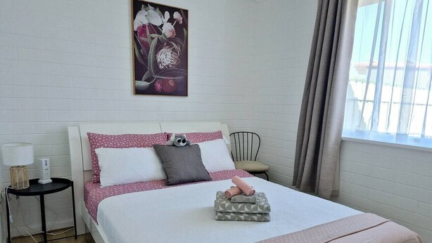 A neatly made bed with pink and white bedding, a small bedside table with a lamp, a chair, and a window with curtains.