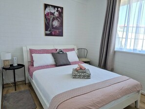 Ein ordentlich bezogenes Bett mit rosa und weißer Bettwäsche, ein kleiner Nachttisch mit Lampe, ein Sessel und ein Fenster mit Vorhängen.