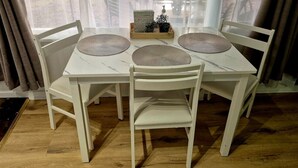 A white dining table with three chairs, a wooden tray with a plant, and a window with curtains.