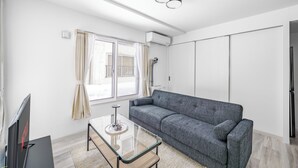 Una sala de estar moderna con un sofá gris, una mesa de centro de vidrio, un televisor de pantalla plana y un amplio ventanal con cortinas.