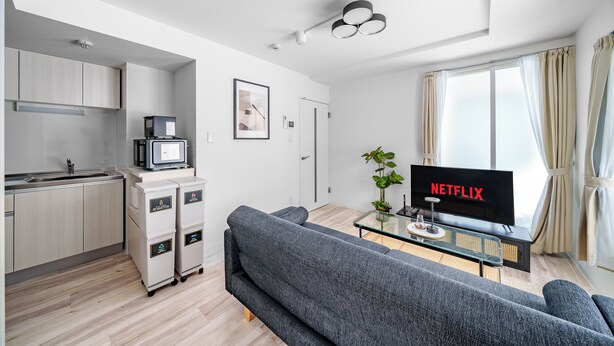 Un apartamento moderno con cocina, un sofá gris, un televisor que muestra 'NETFLIX' y un ventanal con cortinas.