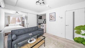 Una habitación moderna y luminosa con un sofá gris, una pequeña mesa de centro, una cama, un armario y una zona de cocina al fondo.