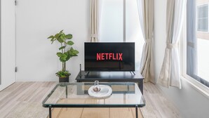 Un salón moderno con una televisión de pantalla plana mostrando 'NETFLIX', una mesa de centro de cristal con un objeto decorativo, una planta en maceta y un ventanal con cortinas.