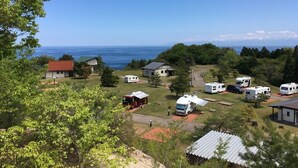 Un camping con vehículos recreativos y casas móviles, una pequeña construcción con techo rojo y vista al océano.