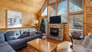 L’intérieur chaleureux d’un chalet avec une cheminée en pierre, une grande fenêtre et un téléviseur posé sur une étagère en pierre.