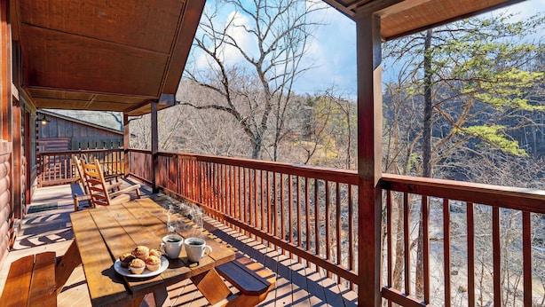 Une terrasse en bois avec une table dressée pour le petit-déjeuner, une balustrade en bois et une vue sur les arbres et les collines.
