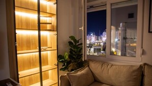 A room with a wooden shelving unit illuminated from within, a beige sofa, and a view of a cityscape at night through a window. - Stone Bridge Apartment (Skopje)