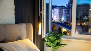A hotel room with a bed, a window with a view of a cityscape, and a plant beside the window. - Stone Bridge Apartment (Skopje)