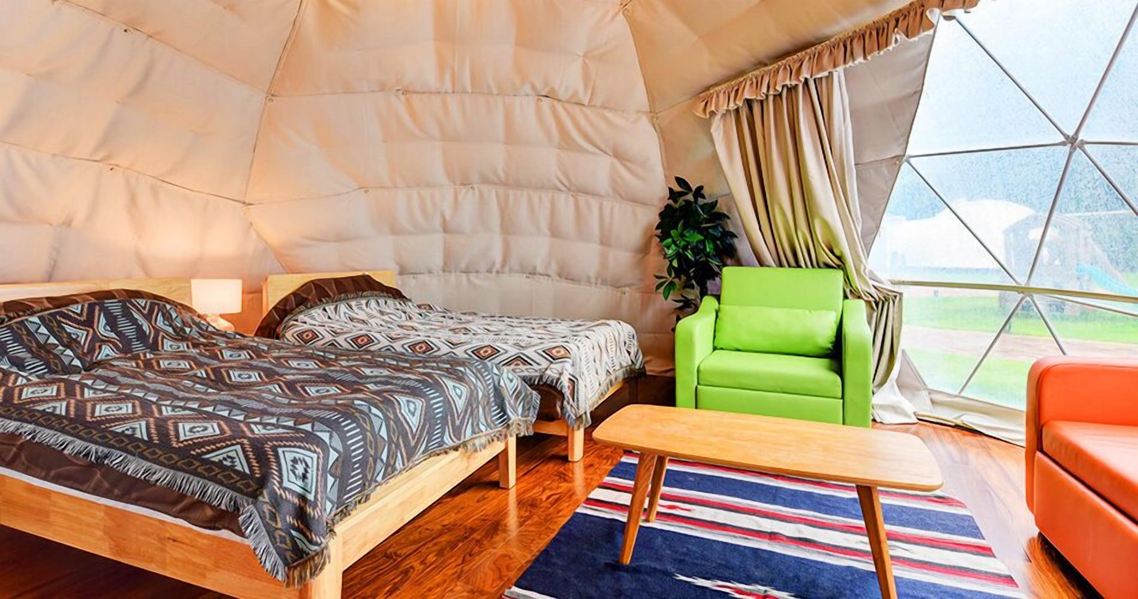 Una habitación tipo tienda de campaña con cama, una silla verde, una mesita y vistas a través de una ventana.