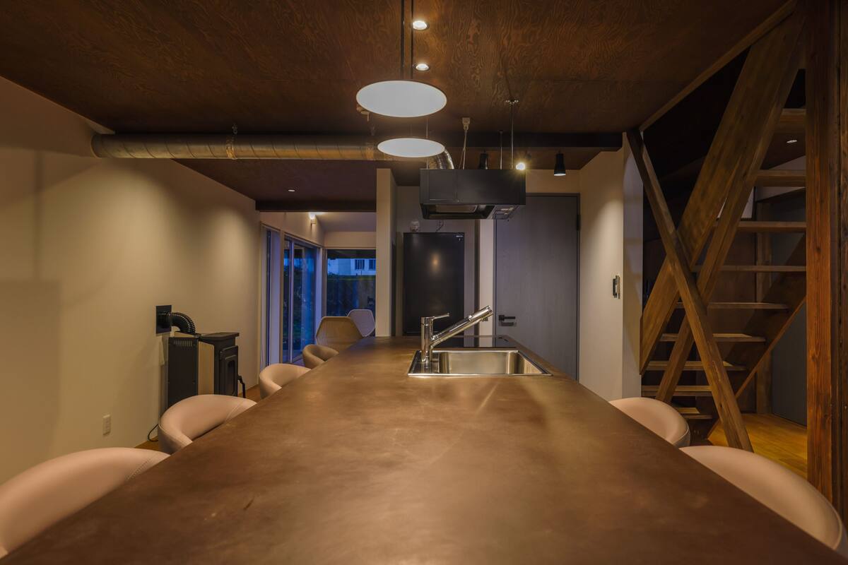 A long conference table with a modern kitchen area, including a sink and cabinets, in a room with wooden beams and a staircase.