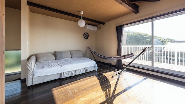 Una habitación con piso de madera, un sofá, una hamaca, un ventilador de techo y un amplio ventanal con vistas al verdor.