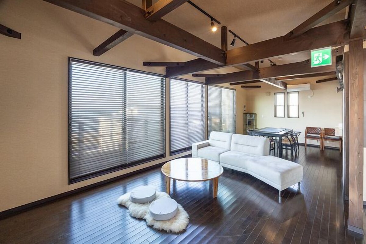 Un vaste espace de vie comprenant un canapé blanc, une table basse ronde et une table à manger avec des chaises. De grandes fenêtres équipées de stores se trouvent à gauche, et un plafond en bois avec des poutres apparentes est visible.