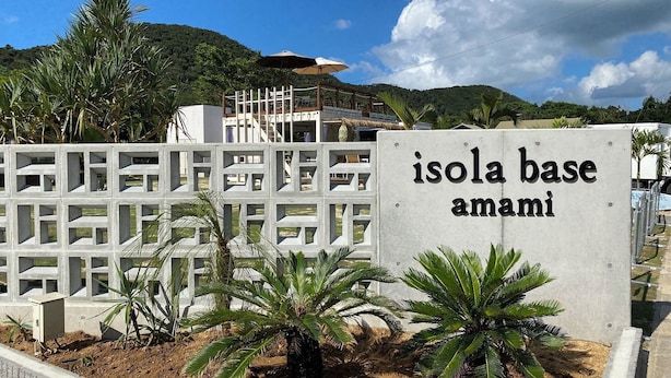A sign for "Isola Base Amami" with a concrete structure and palm trees in the foreground. - Isola Base Amami (Tatsugo)