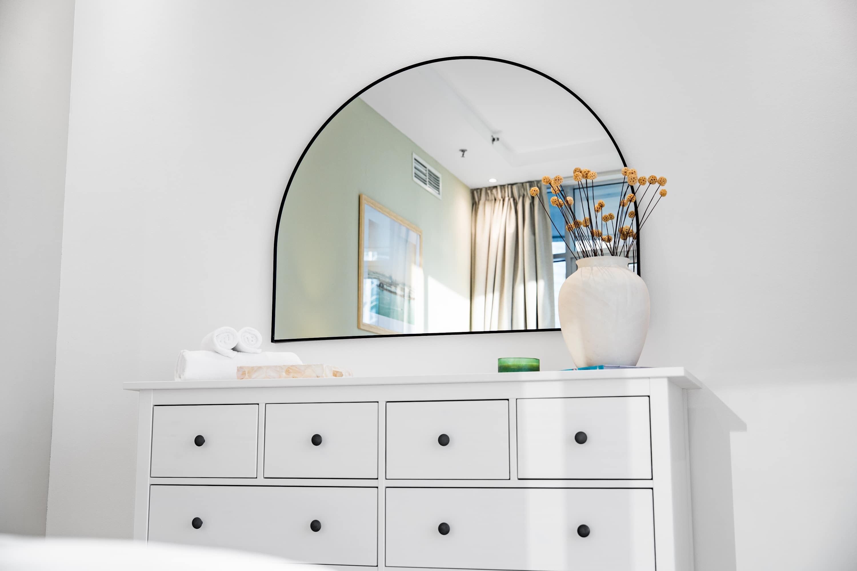 A white dresser with a mirror above it, a vase with flowers, and a window with curtains.
