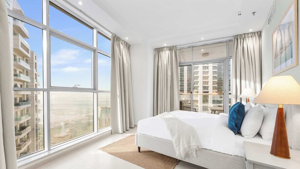 Una habitación de hotel moderna con una cama grande, una mesita de noche con lámpara y vistas de la ciudad a través de ventanales de gran altura.