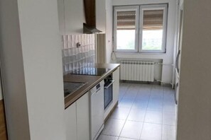 A kitchen with white cabinets, a stove, and a window with blinds. - Skopje City Apartment 1 (Skopje)