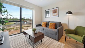 Uma sala de estar moderna com um sofá cinzento, uma poltrona verde, uma mesa de centro de madeira e uma grande janela com vista para a paisagem urbana.