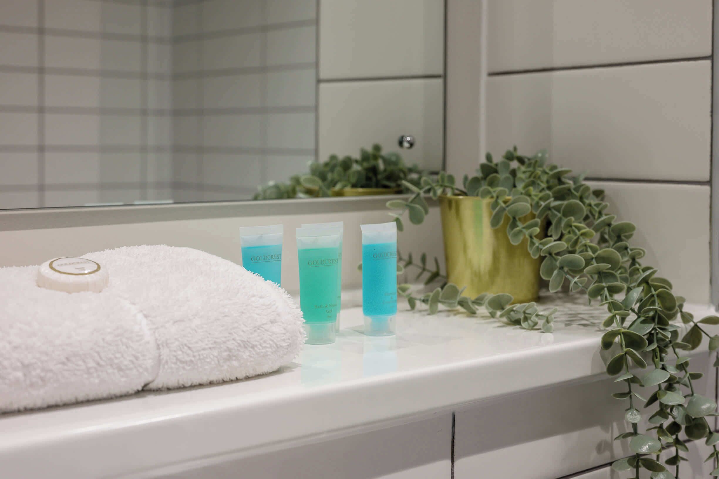 A bathroom with a white towel, three Goldcrest bath and shower gel bottles, and a potted plant on the counter.