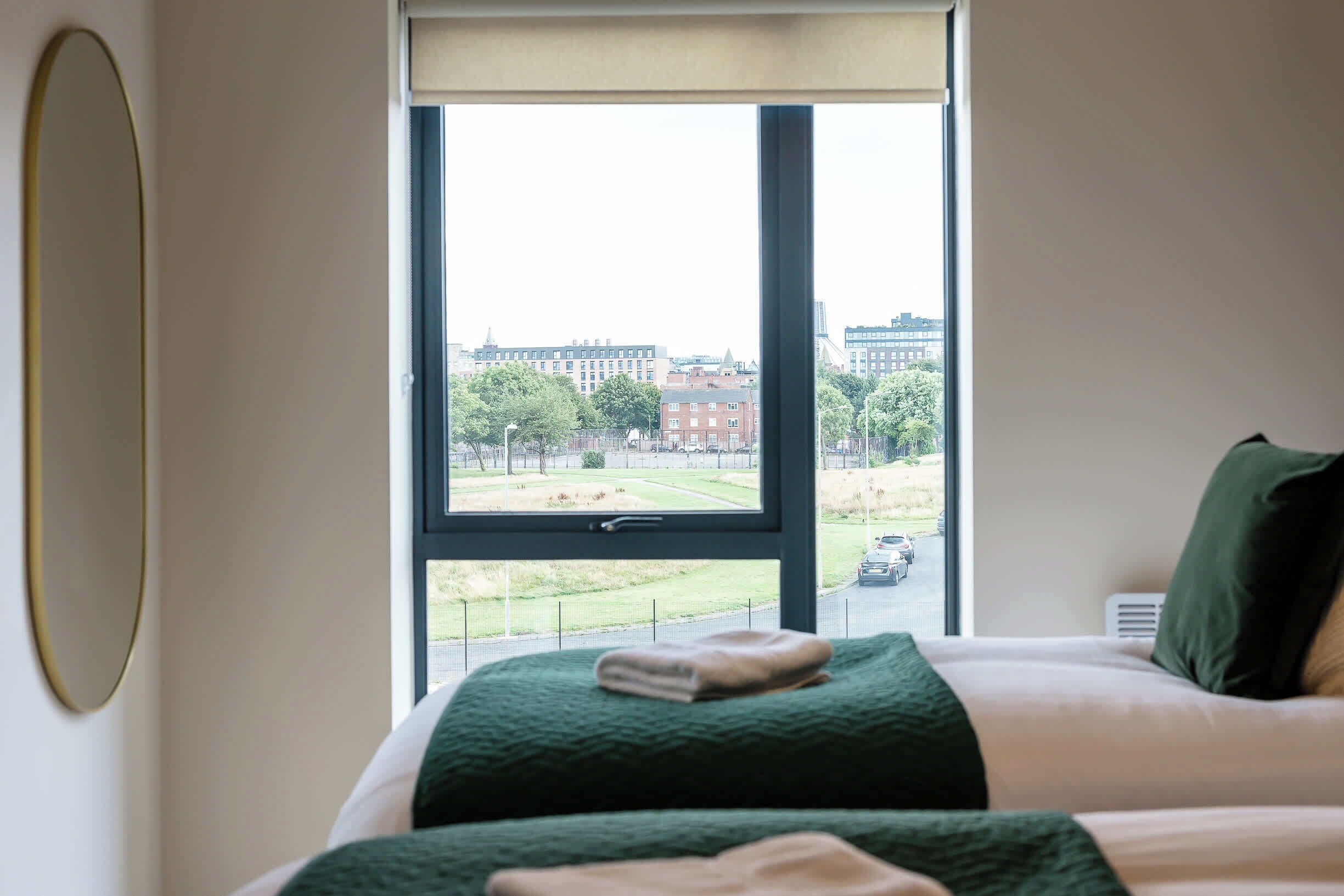 A hotel room with a large window, a bed with green bedding, and a view of a cityscape.