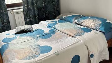 Un dormitorio con una cama cubierta por una colcha celeste y blanca a lunares, una ventana con cortinas y un radiador.