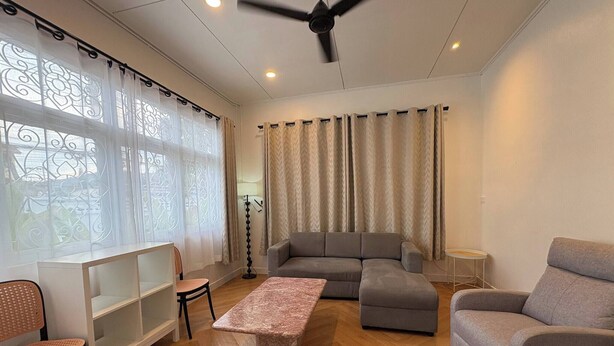A modern living room with a grey sectional sofa, a small table, and a bench. There is a ceiling fan, a floor lamp, and a window with decorative curtains. - 3BR Family Villa - 1 km to Chalong Pier with WiFi (Chalong)