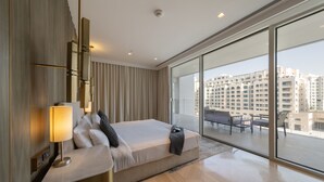 Ein modernes Hotelzimmer mit einem großen Bett, einem Nachttisch mit Lampe, einer Couch und einem Balkon mit Blick auf hohe Gebäude.