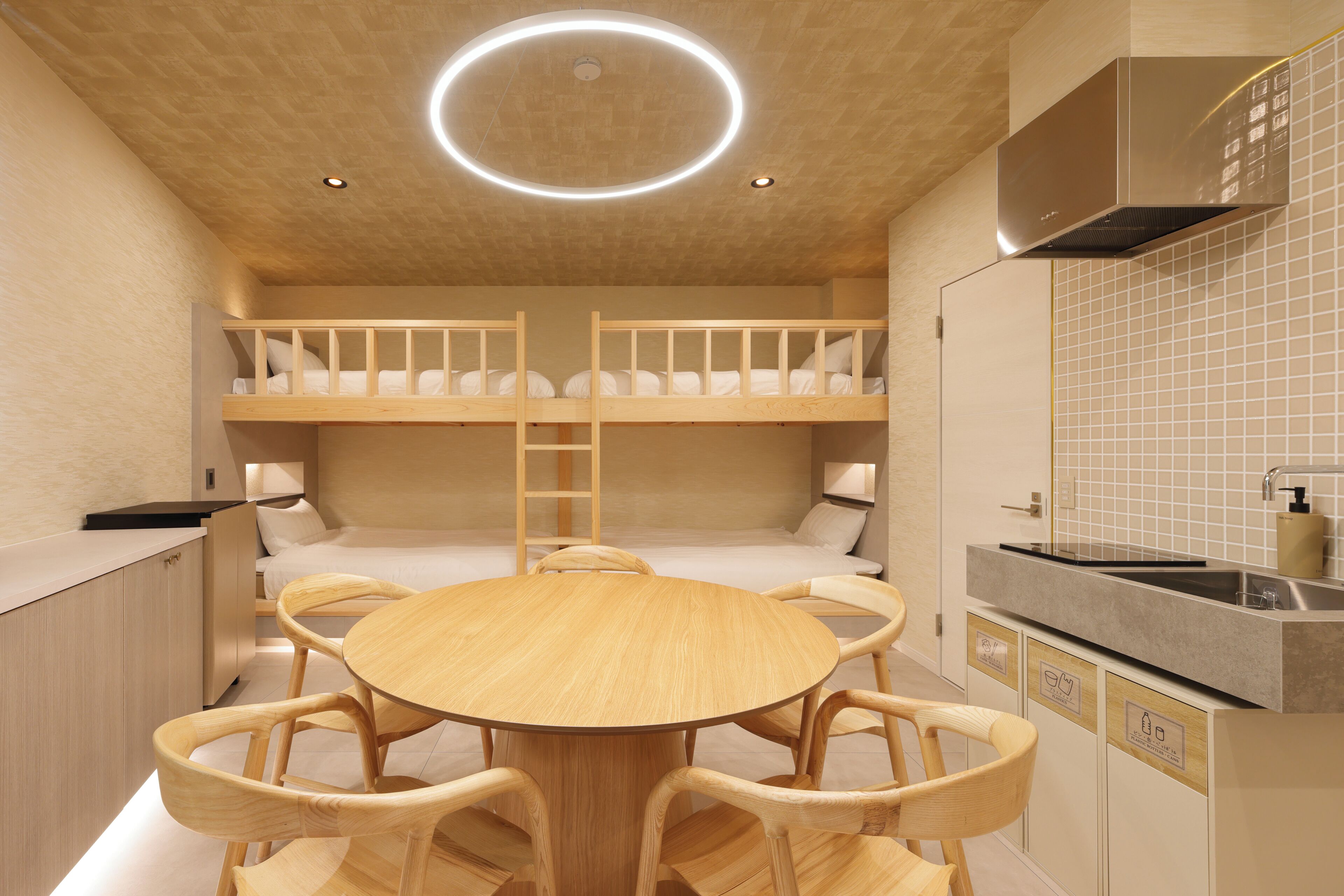 A bunk bed room with a round wooden table and chairs, a kitchen area with a sink and cabinets, and a ceiling with recessed lighting.