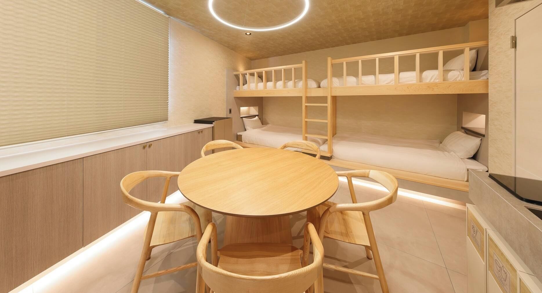 A bunk bed room with a round wooden table and chairs, a window with blinds, and a ceiling light.