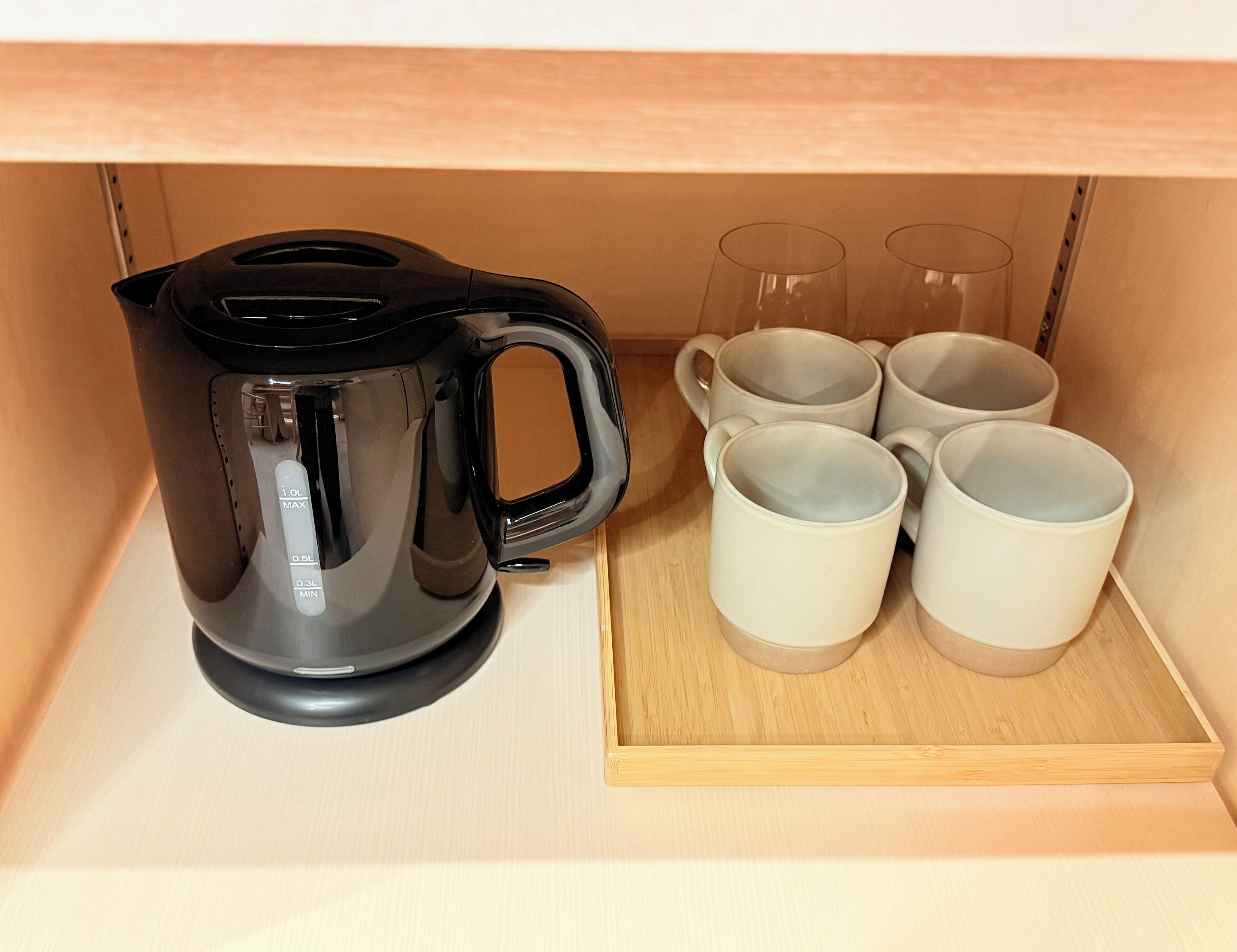 A black coffee maker and four white cups on a wooden tray in a kitchen cabinet.