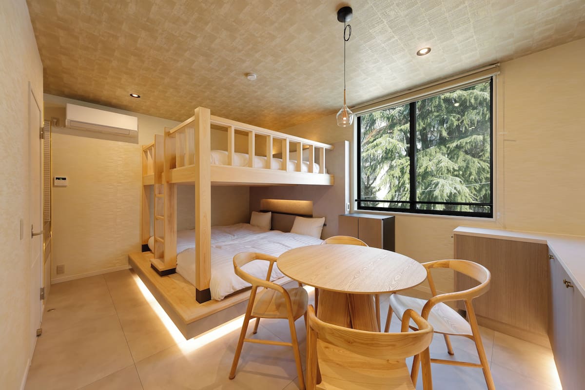 Une chambre avec un lit superposé, une table ronde en bois avec des chaises, une grande fenêtre donnant sur les arbres et un plafond doté d’un éclairage encastré.