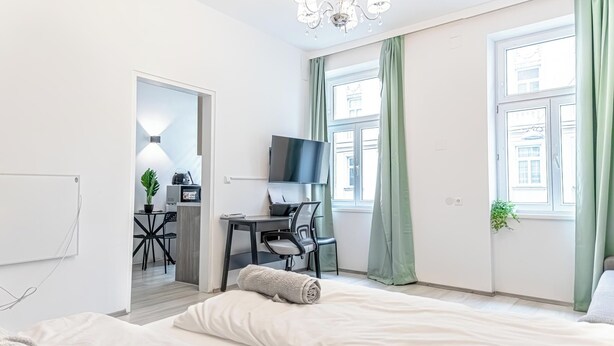 Ein modernes Hotelzimmer mit einem Bett, einem Schreibtisch mit Computer, einem Fernseher und Blick auf die Stadt durch große Fenster.