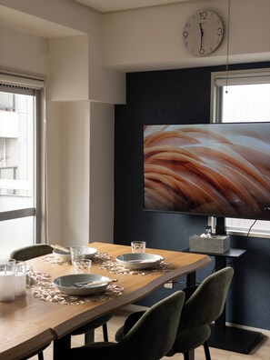 Een eetruimte met een houten tafel gedekt voor twee personen, een grote flatscreen-televisie aan de muur bevestigd, en een raam dat voor natuurlijk licht zorgt.