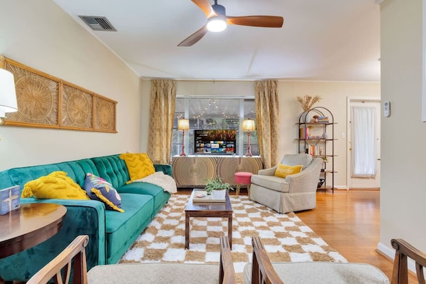 A living room with a teal sofa, a grey armchair, and a coffee table. There is a ceiling fan, a TV, and a bookshelf.