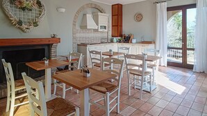 Zona pranzo con sedie e tavoli bianchi, un caminetto, una cucina con mobili bianchi e vista attraverso una porta in vetro.