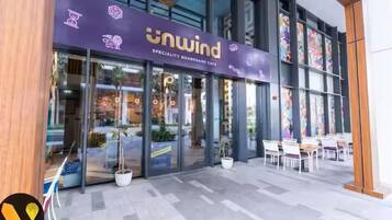 Das äußere Erscheinungsbild eines Brettspielcafés namens 'Unwind' mit Glasfassade, Außenbereich mit Sitzgelegenheiten und einem modernen architektonischen Design.