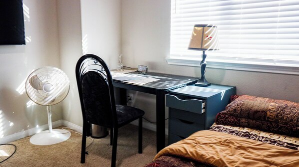 Une chambre comprenant un bureau, une chaise, un lit et un ventilateur sur pied.