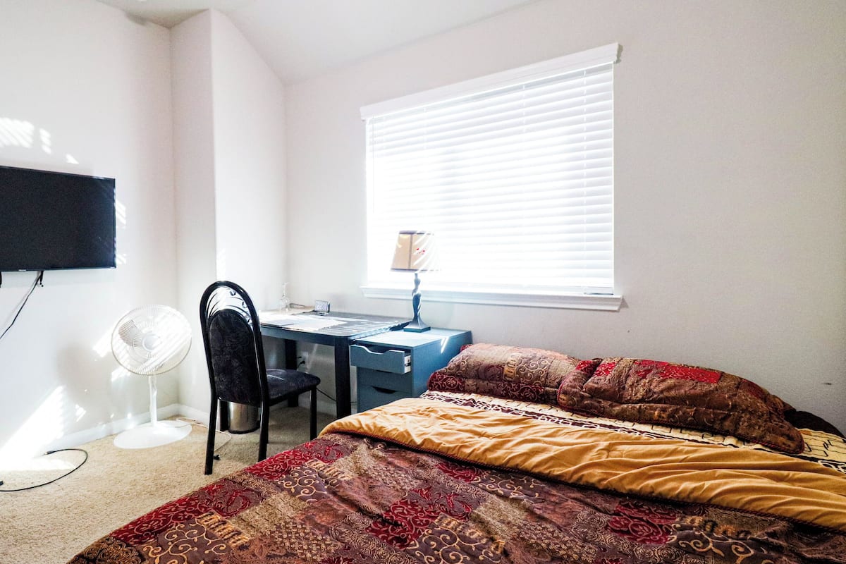 A bedroom with a desk, chair, bed, and a standing fan.